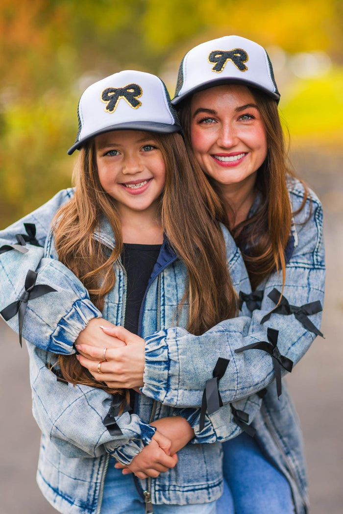 Mom & Me - Bow Denim Jacket - CHILD-3XS-6-12 Months - Mother and daughter wearing matching light blue bow-accented denim jackets and white hats with black bow designs outdoors.