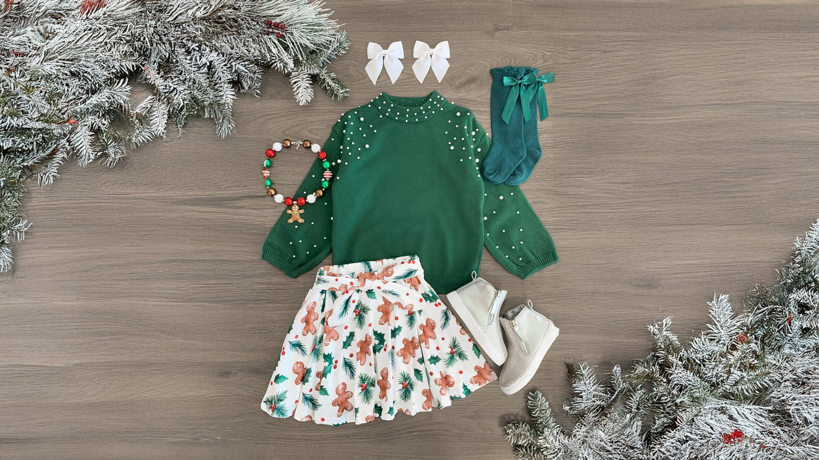 Girls' holiday outfit featuring a green long sleeve top with pearl details and a white skirt adorned with a gingerbread and holly print. The skirt has a stretchy waistband for comfort, and the ultra-soft fabric provides all-day ease. Perfect for the holiday season, this set offers a festive, stylish look that can be paired with booties or a matching bow.