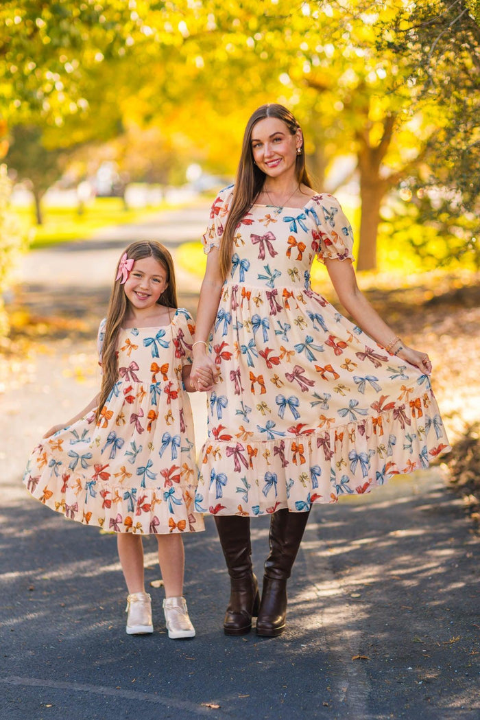 Mom & Me - Peachy Pink Bow Dress - CHILD-3XS-6-12 Months - Matching cream dresses with multi-colored bow prints, pleated skirt, and short sleeves, worn by mother and daughter outdoors.