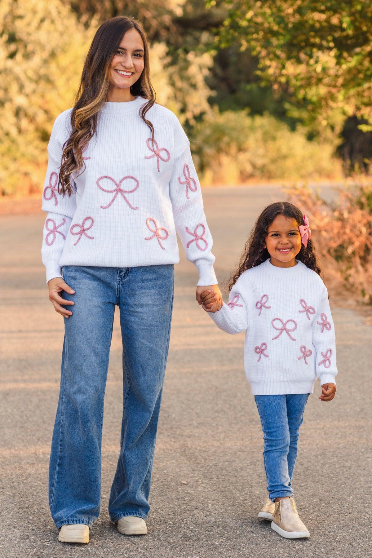 Woman and young girl holding hands, both wearing matching white sweaters with pink bows outdoors.