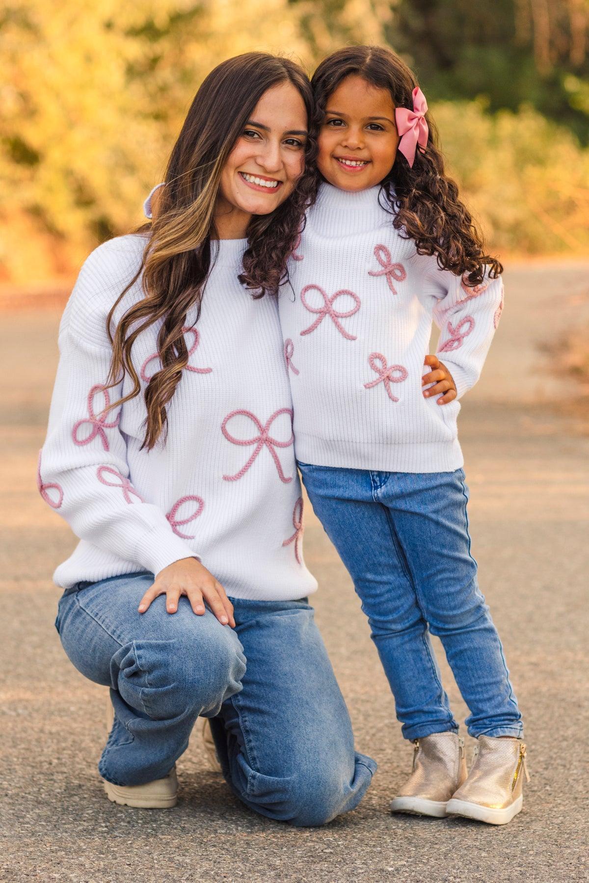 Woman and child wearing matching sweaters with pink bows outdoors.