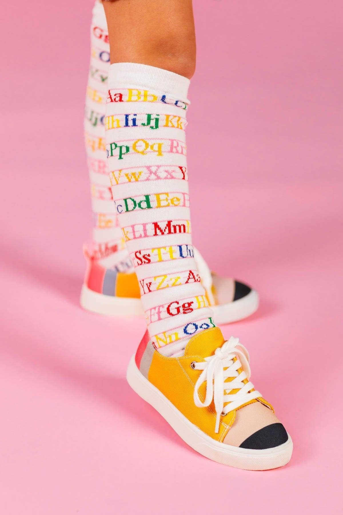 Pencil Sneakers - KIDS-8T - Yellow canvas sneakers with white laces and black toe caps worn with colorful alphabet socks against a pink background.