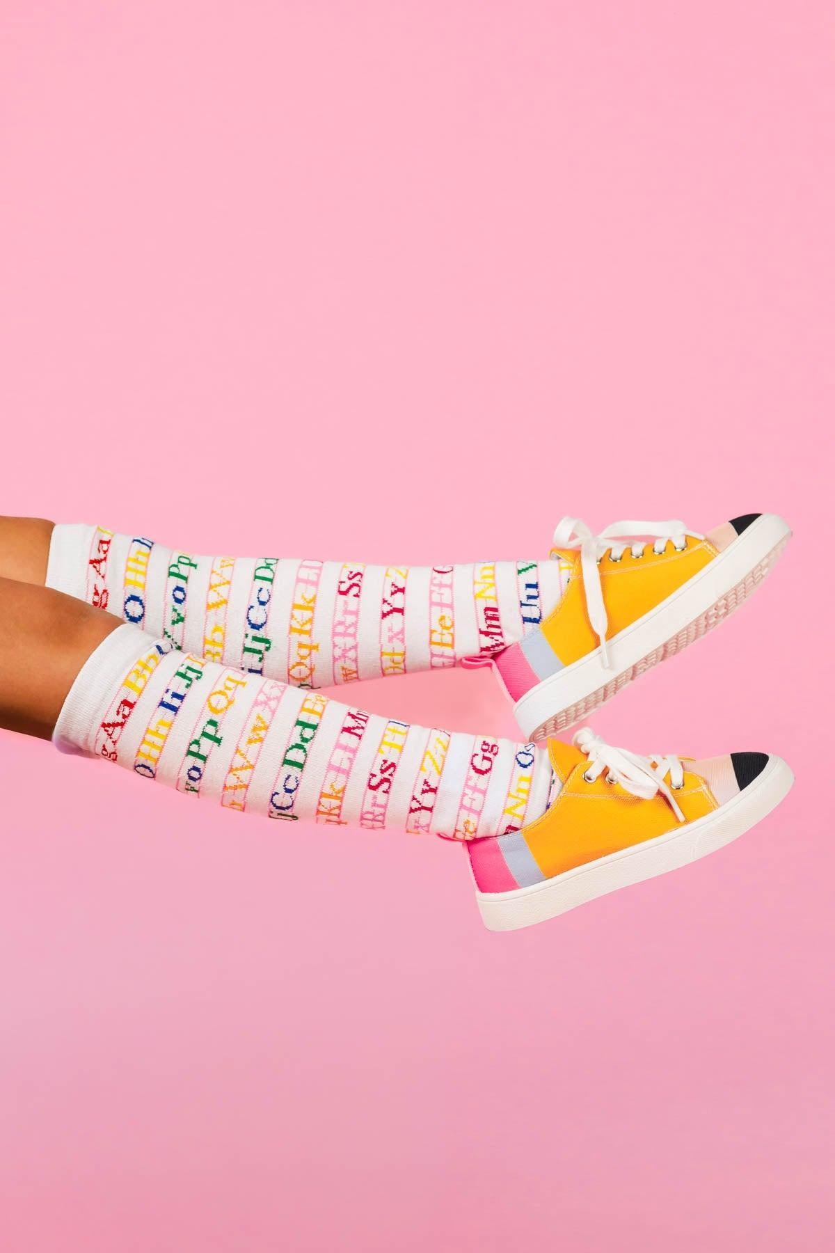 White Alphabet Knee High Socks - Default Title - White knee-high socks with colorful alphabet print worn with yellow and pink canvas sneakers against a pink background.