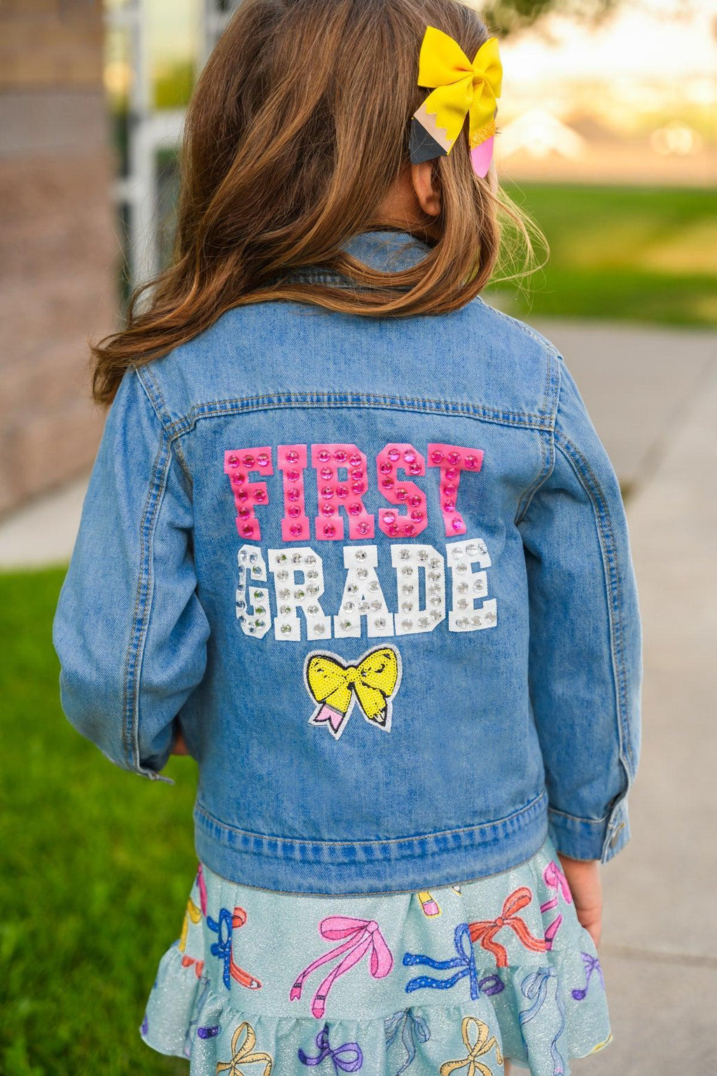 "Preschool - Sixth Grade" Denim Jacket - PRESCHOOL / 3T - Light blue denim jacket for kids with pink, white, and yellow "First Grade" text and a yellow bow on the back.