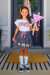 "Pre-K - Sixth Grade" Notebook Skirt Set - PRE-K / 2T - Girl wearing black skirt with colorful school-themed notebook print, white "First" ruffle sleeve top, striped socks, and.