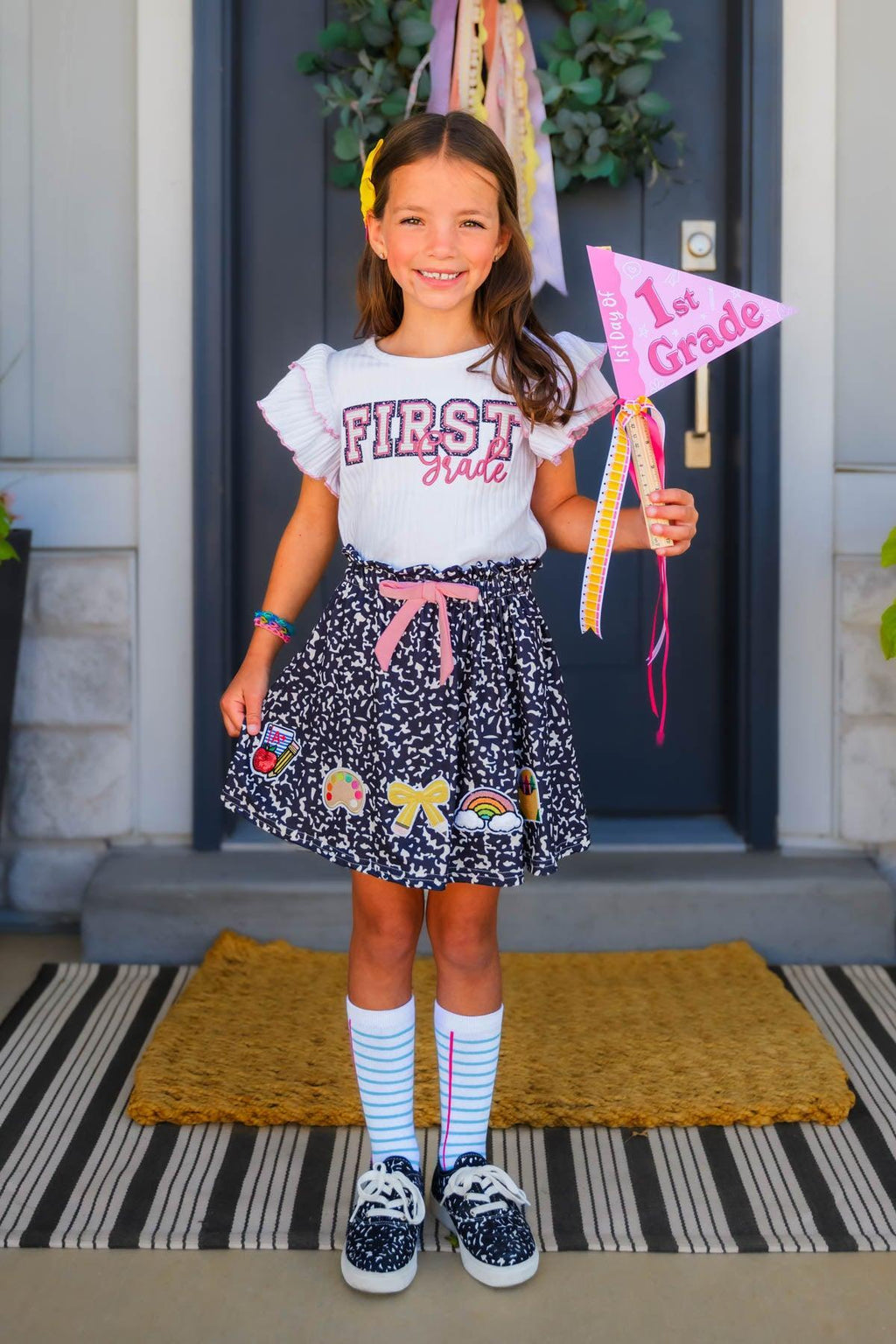 "Pre-K - Sixth Grade" Notebook Skirt Set - PRE-K / 2T - Girl wearing black skirt with colorful school-themed notebook print, white "First" ruffle sleeve top, striped socks, and.