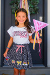 "Pre-K - Sixth Grade" Notebook Skirt Set - PRE-K / 2T - Girl wearing black notebook print skirt with pink bow and white ruffle-sleeve First Grade shirt holding a pink 1st Grade flag.