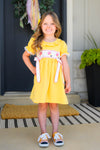 Yellow Pencil & Bows Dress - 3T - Yellow short-sleeve dress with pencil and bow appliques, ruffled sleeves, and white accents, worn by a smiling girl.