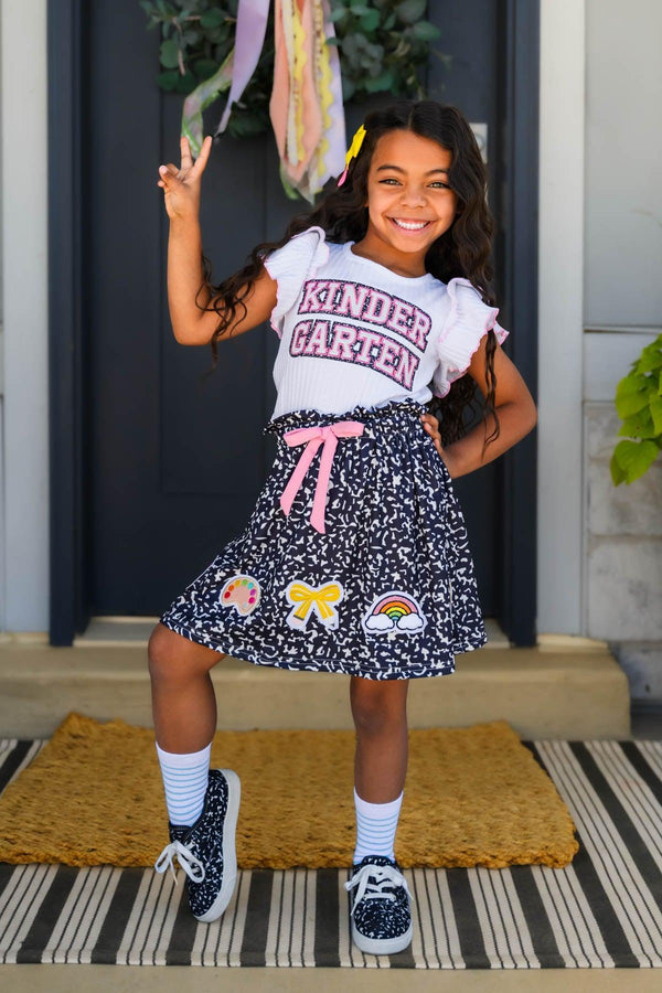 "Pre-K - Sixth Grade" Notebook Skirt Set - PRE-K / 2T - Girl wearing black notebook print skirt with colorful designs and white "Kindergarten" ruffle sleeve t-shirt.