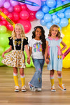 "Pre-K - Sixth Grade" Bow Denim Pant Set - PRE-K / 2T - Three young girls wearing colorful outfits, including denim pants with a bow detail, standing against a bright balloon.