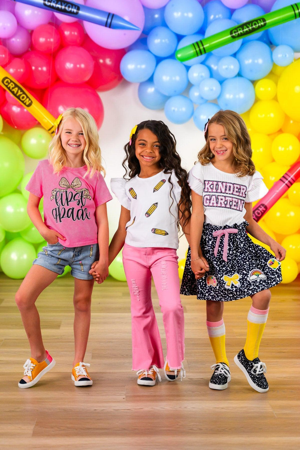 "Pre-K - Sixth Grade" Notebook Skirt Set - PRE-K / 2T - Three girls standing in front of colorful balloon wall; left girl wears pink graphic t-shirt with denim shorts, middle girl.