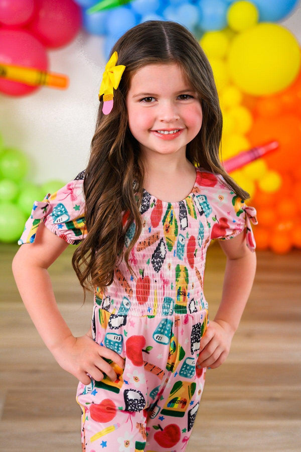 Pink School Supplies Jumpsuit - 3T - Pink short-sleeve jumpsuit with colorful school supplies print worn by smiling girl with long hair and yellow hair clip.