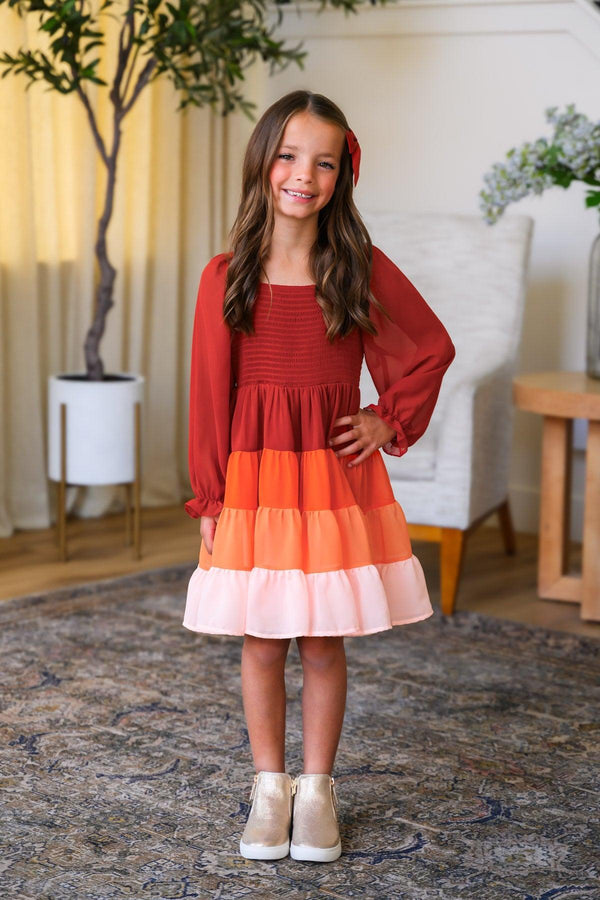 Mom & Me - Fall Ruffle Ombre Dress - CHILD-6-12 Months - Long-sleeve dress with tiered ruffle skirt in rust, orange, and pink ombre colors, worn by a smiling girl.