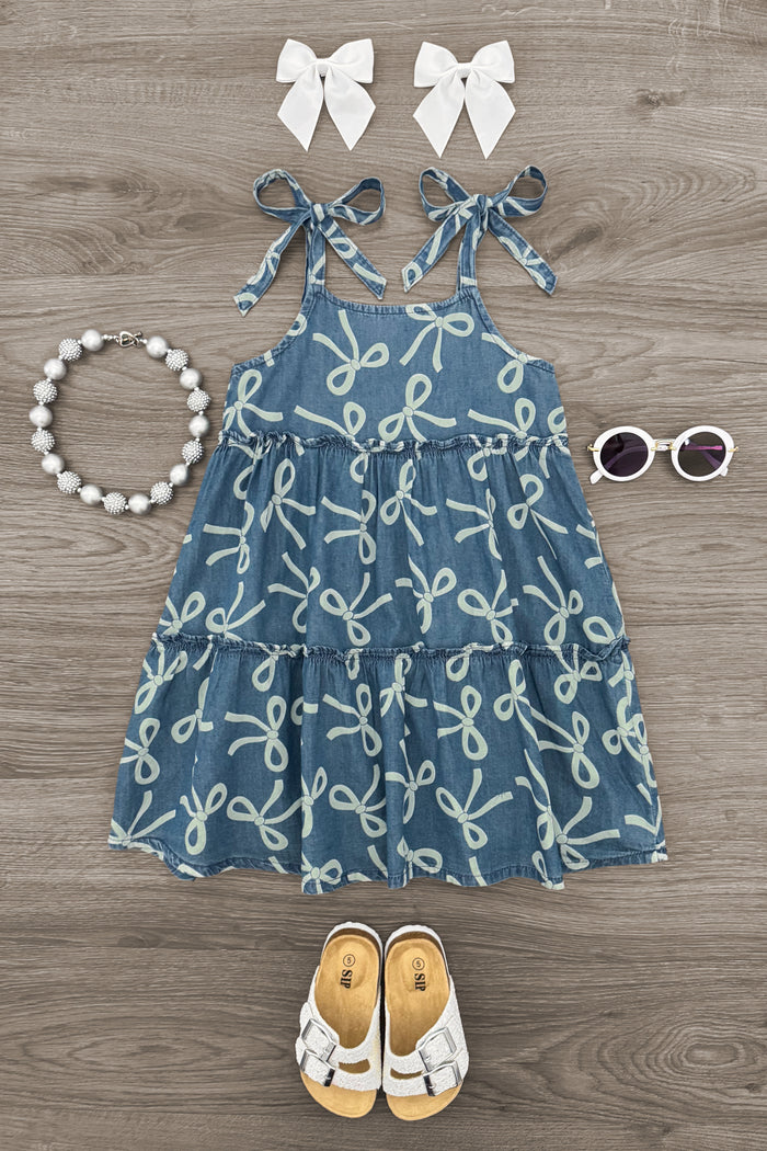Denim Bow Dress - 6-12 Months - Blue denim dress with white bow patterns and tied shoulder straps, paired with white bow hair clips, white sandals, a bead.