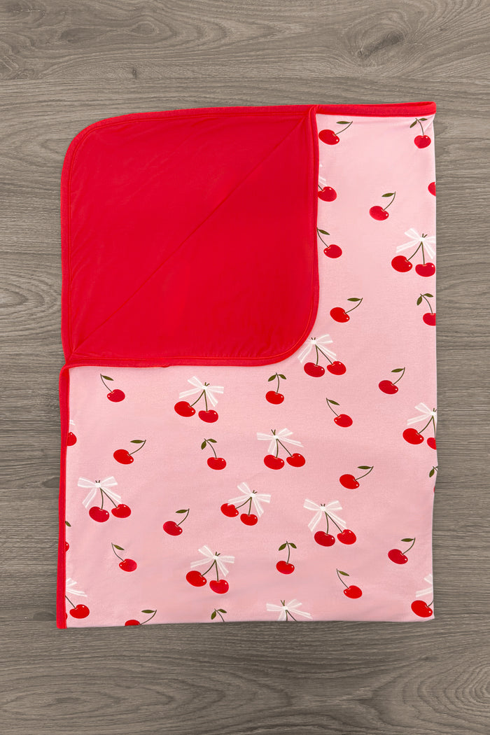 Bamboo Sweet Cherry Double Sided Blanket - Default Title - Pink blanket with red cherry pattern and solid red reverse side on wooden floor.