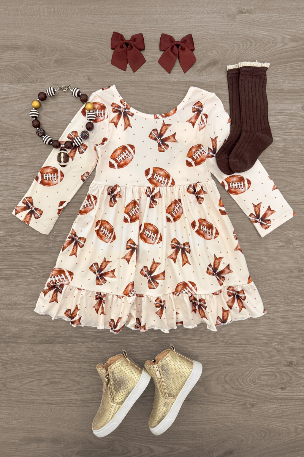 Football Bow Dress - 6-12 Months - Cream long-sleeve dress with football and bow prints, ruffled hem, paired with brown socks, gold sneakers, red bow clips,.