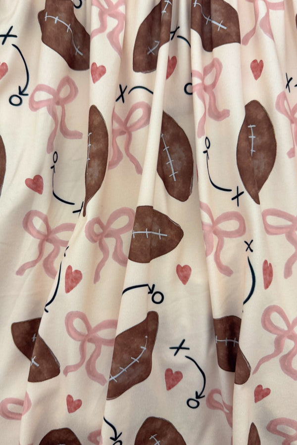 "Game Day" Football Bow Dress - 6-12 Months - Cream fabric dress with brown footballs and pink ribbons, playful sports-themed pattern.