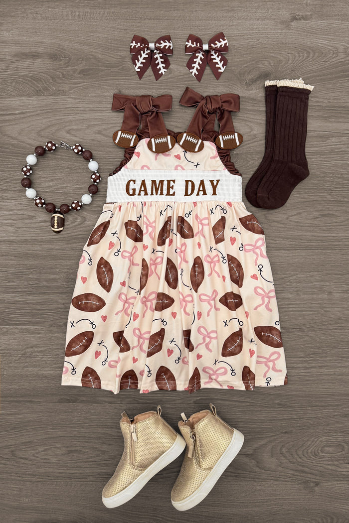 "Game Day" Football Bow Dress - 6-12 Months - Cream dress with brown football and pink bow prints, brown bow straps, paired with brown socks, tan boots, brown bow hair.