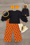 Black & Orange Polka Dot Denim Pant Set - 6-12 Months - Black long-sleeve top paired with orange polka-dot denim pants, knit beanie, orange bows, necklace, and cream boots.
