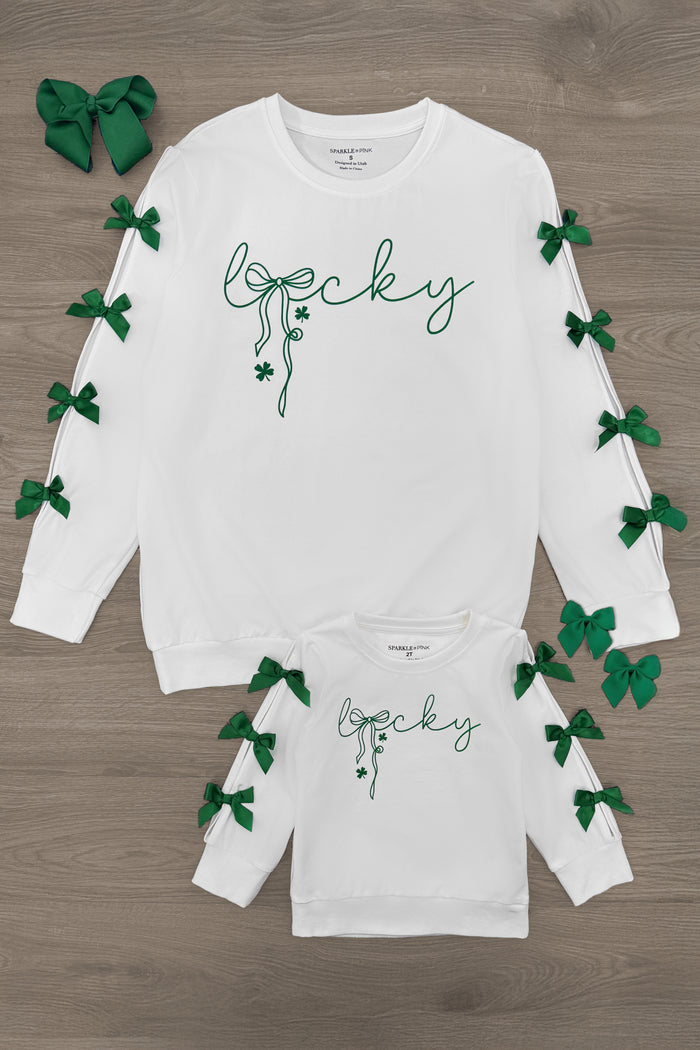Mom & Me - "Lucky" Bow Top - CHILD-6-12 Months - White long-sleeve tops with green “lucky” script, shamrock detail, and green bows along sleeves, adult and child sizes.