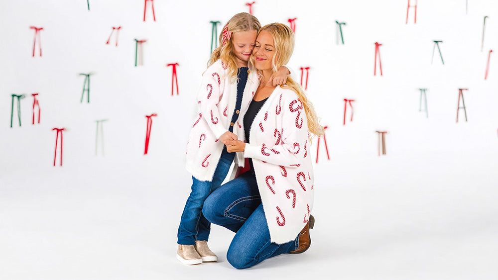 Mother and daughter hugging in matching white candy cane cardigans and jeans against a backdrop of red and green bows.
