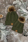 Mom & Me - SIP Colored Beanies - Child / Black - Two olive green knit beanies with faux fur pom-poms and leather SIP logo patches on a snowy wooden surface with pine.