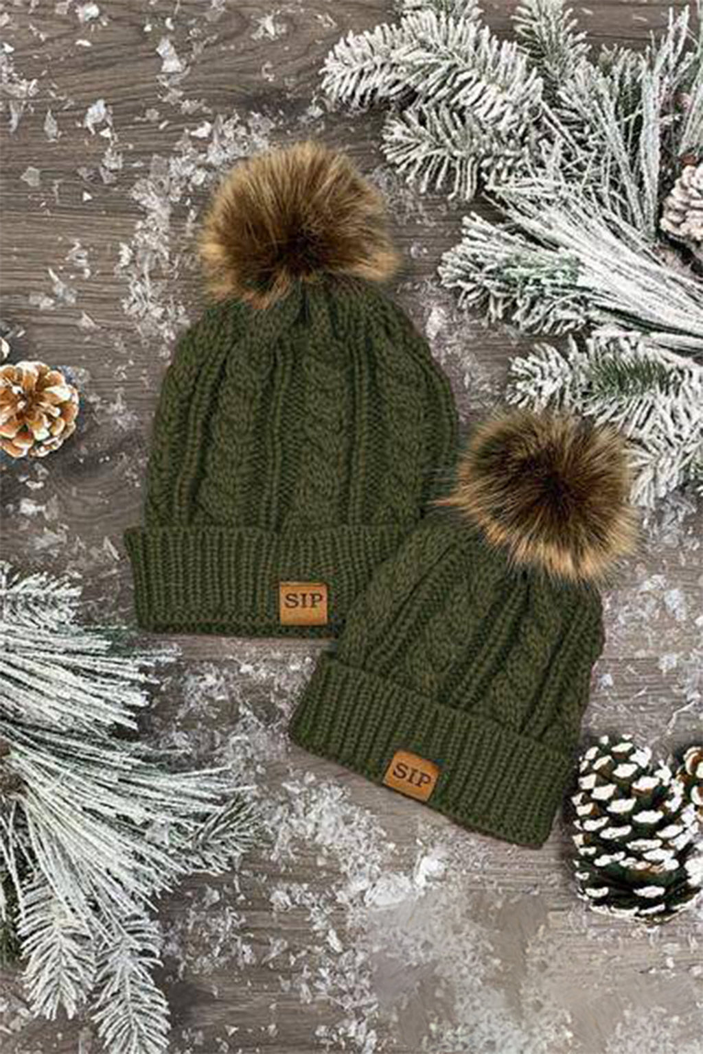 Mom & Me - SIP Colored Beanies - Child / Black - Two olive green knit beanies with faux fur pom-poms and leather SIP logo patches on a snowy wooden surface with pine.