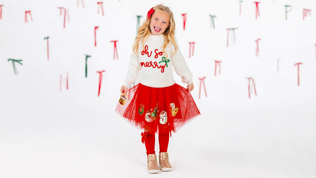 Girl wearing a white “Oh So Merry” sweater, red tulle skirt with Christmas appliqués, red leggings, and gold boots.