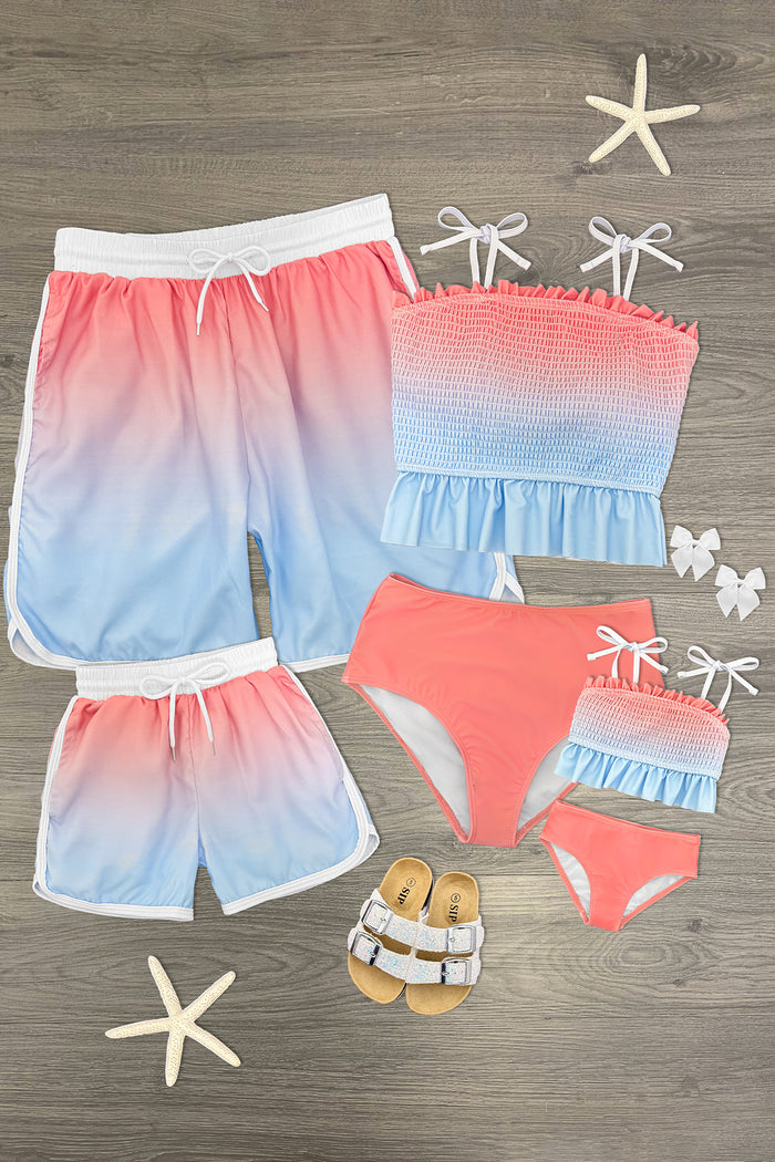 Pink and blue ombre family swimsuits set with shorts, bandeau top, bikini, and toddler swimwear on wood surface.