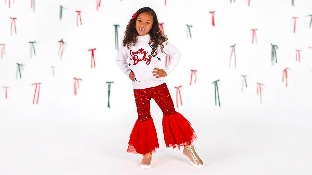 Girl wearing a white “Santa Baby” sweater, red sequin bell-bottom pants with tulle flares, and gold boots against a backdrop of red and green bows.