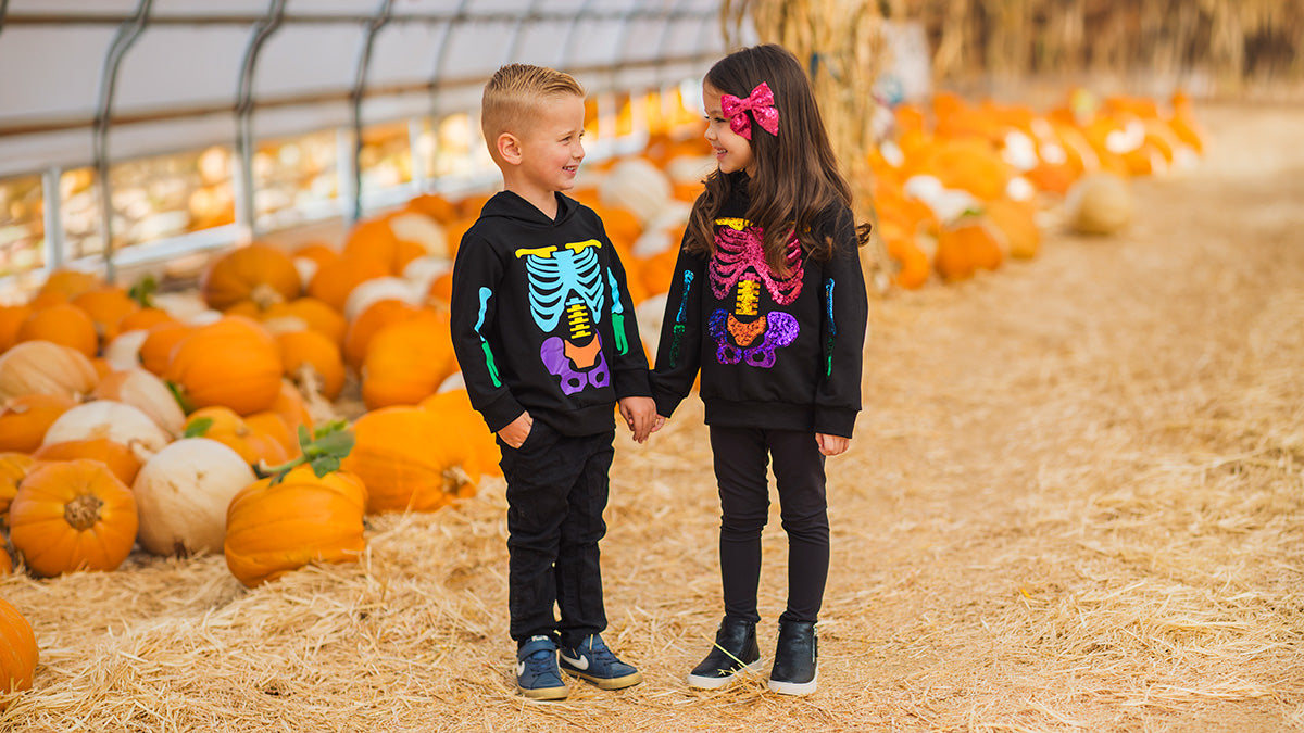 Boy and girl holding hands in a pumpkin patch while wearing matching black Halloween hoodies with colorful skeleton designs.