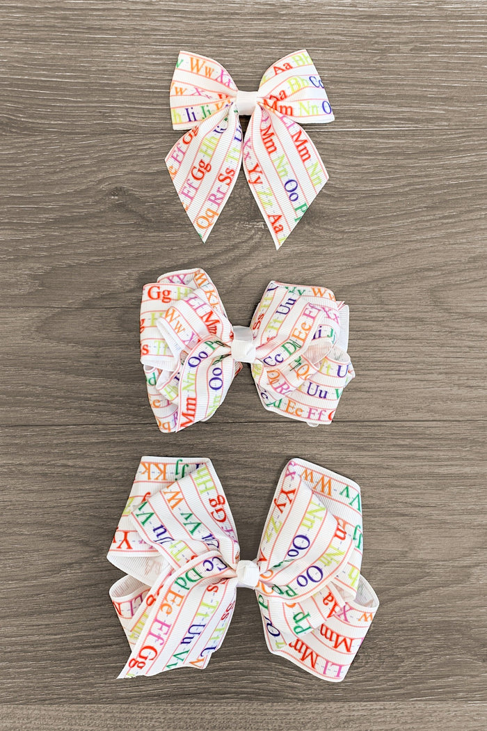 Alphabet Bows - 3 inches - Three white hair bows with colorful alphabet letters in small, medium, and large sizes on a wooden surface.