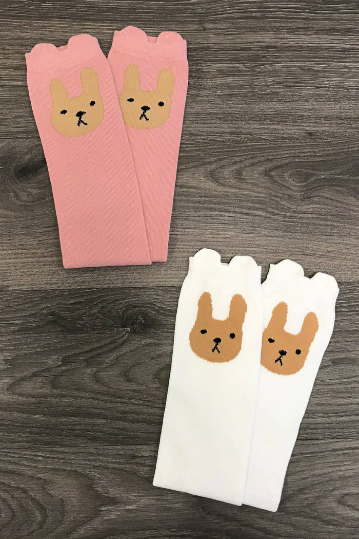 Bashful Bunny Socks - White - Pink knee-high socks with bunny faces and scalloped edges beside white knee-high socks with brown bunny faces on a dark wood .