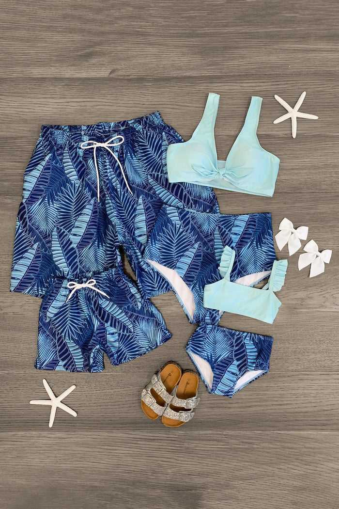Blue tropical leaf print swim trunks for men and blue tropical leaf print bikinis with light blue tops for women and girls.